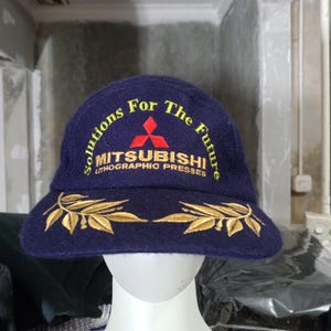 May include: A navy blue baseball cap with gold embroidered leaf designs on the brim. The cap features the text "Solutions For The Future" and "MITSUBISHI LITHOGRAPHIC PRESSES" in gold lettering and a red logo.