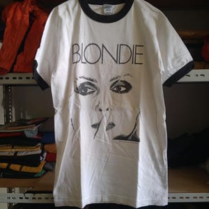 May include: White ringer t-shirt featuring the word "BLONDIE" in black block letters above a black and white graphic of a woman's face. The shirt has a black collar and black sleeve trim.
