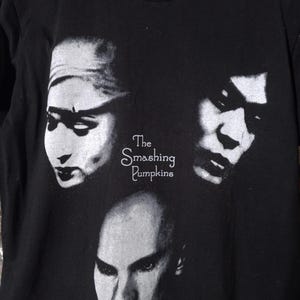 May include: A black short-sleeved t-shirt featuring a white graphic print of three faces and the text The Smashing Pumpkins in the center. The design is monochromatic, showcasing high-contrast portraits of individuals with distinct facial features.