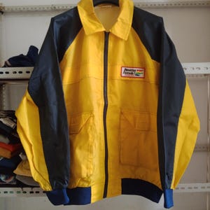 May include: A yellow and navy blue zip-up jacket with a collar and two front pockets. The jacket features the Benetton Formula 1 Racing Team logo on the chest. The jacket has a ribbed hem and cuffs.