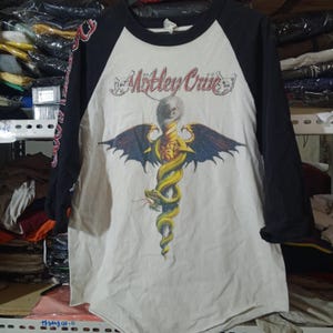 以下が含まれることがあります： 黒の七分袖が付いた白いベースボールスタイルのTシャツ。前面には、ダークブルーの翼と頭蓋骨を持つ短剣に巻き付いた黄色いヘビのグラフィックが描かれており、その上には赤い文字でMötley Crüeとプリントされています。