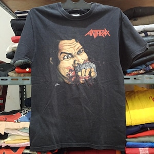 Puede incluir: Camiseta negra de la banda Anthrax con un gráfico de una cara con un puño apretado en la boca, sangre y el nombre de la banda en rojo. La camiseta está colgada de una percha negra.