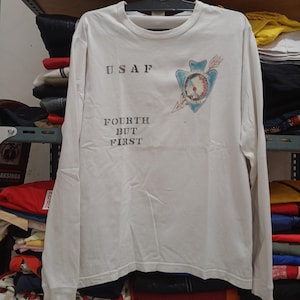 May include: White long-sleeve shirt with the text "USAF" above "FOURTH BUT FIRST". A blue and red graphic is on the right chest. The shirt is made of a soft, comfortable material.