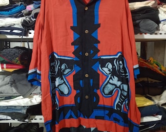 May include: A red shirt with a distinctive pattern. It has black and blue geometric shapes and two elephant figures. The sleeves have blue detailing. The shirt has a collar and wooden buttons. The shirt is a button-down style.