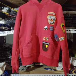 May include: A red long-sleeve hooded sweatshirt featuring a front kangaroo pocket and various embroidered patches, including text that reads CANADA, Dsquared, 1964, and Dsquared Brotherhood, with a ribbed V-neck detail at the collar.
