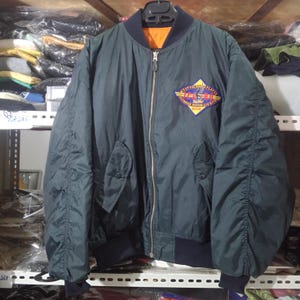 May include: A dark green bomber jacket with a front zipper closure, ribbed collar, and cuffs. The jacket features a diamond-shaped patch on the left chest with the text "MOST VALUABLE PLAYER" and "JAPANESE RECORD LTD HITS" in yellow and red.