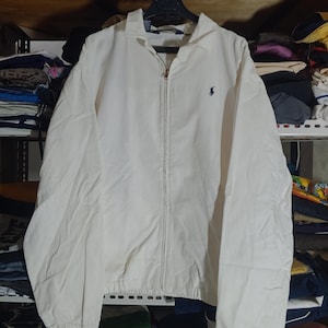 Puede incluir: Chaqueta con cremallera de color crema con cuello y un pequeño logotipo azul marino. La chaqueta tiene una cremallera completa y un ajuste relajado. La tela parece ligera, adecuada para usar en capas.