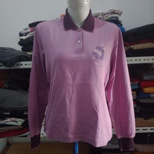 Puede incluir: Un polo de manga larga de color lila claro con cuello y puños morados oscuros. La camisa presenta un diseño de bordado floral en el pecho izquierdo y una tapeta de tres botones. Los puños tienen un borde festoneado.