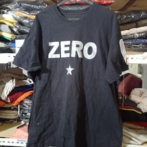 May include: A black short-sleeve t-shirt featuring the word ZERO printed in large white capital letters across the chest, with a small white five-pointed star graphic positioned directly below the text and a white circular logo on the left sleeve.