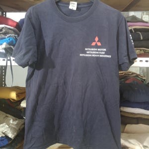 May include: Navy blue t-shirt with a crew neck and short sleeves. The shirt features the Mitsubishi Motors logo in red, along with the text "MITSUBISHI MOTORS, MITSUBISHI FUSO, MITSUBISHI HEAVY INDUSTRIES" in white.
