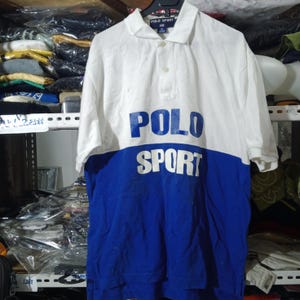 May include: A short-sleeve polo shirt featuring a color-blocked design with a white upper section and a blue lower section. The text POLO SPORT is printed across the chest in blue on the white portion and white on the blue portion.