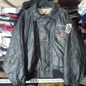 May include: A black jacket with a collar and a zipped chest pocket. The jacket features a patch with "TRD" text and a chequered flag design. It has white stitching and a black ribbed hem and cuffs.