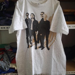 Puede incluir: Camiseta gris claro con un gráfico en blanco y negro de una banda. Los miembros de la banda están representados de pie, con un diseño beige en el fondo. La camiseta tiene mangas cortas y cuello redondo.