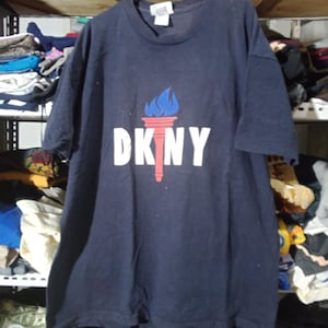 May include: Navy blue DKNY t-shirt with a torch graphic, blue flames, and a red handle. The white DKNY logo is below the torch. The shirt has a crew neck and short sleeves. A casual top.