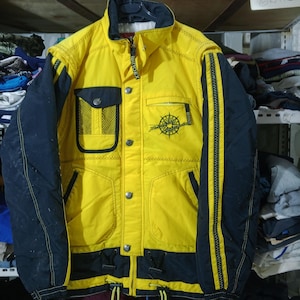May include: Yellow and navy blue winter jacket with the text "Windex" on the zipper and sleeve. The jacket has a compass design on the front and a mesh pocket. The jacket has a drawstring at the bottom.