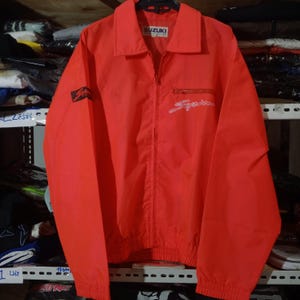 May include: A bright orange windbreaker jacket featuring a full-length front zipper, a classic collar, and elastic cuffs and hem. The Suzuki logo is printed on the inner neck label, with a script logo on the left chest pocket and right sleeve.