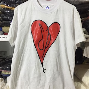 May include: A white short-sleeve t-shirt featuring a large red heart graphic on the chest. The heart is outlined in black and contains a black stylized script logo inside. The shirt has a crew neckline and is displayed on a black plastic hanger.