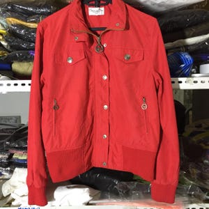 May include: A bright red jacket featuring a full-length front zipper, a snap-button placket, and two chest pockets with snap closures. The jacket includes two zippered side pockets with circular logo pulls, ribbed cuffs, and a ribbed hemline.