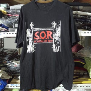 May include: A black short-sleeved t-shirt featuring a graphic print with the text "MAKE YOUR LIFE IN DISORDER S.O.R SCANDAL OF RIOT" in red and white. The design includes two stylised skeletal figures on the sides holding the central text block.