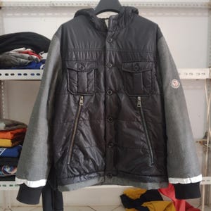 Puede incluir: Chaqueta negra acolchada con capucha y mangas grises. La chaqueta tiene dos bolsillos en el pecho, dos bolsillos inferiores con cremalleras y una botonadura frontal. Un logotipo es visible en la manga izquierda. La chaqueta está colgada en una percha negra.