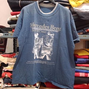 May include: Blue short-sleeved t-shirt with a gray neckline. The shirt features the text "Mercedes Benz" above a graphic of two figures. Below the graphic is the text "Self ID: 3045" and additional text.