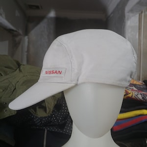 May include: A white baseball cap with a curved peak. The cap has the red "NISSAN" logo on the front left. The cap is made of a light-coloured fabric and is displayed on a mannequin head.