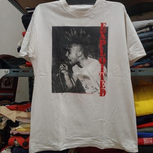 Pode incluir: Camiseta branca com uma imagem em preto e branco de uma pessoa cantando em um microfone com um penteado moicano. A palavra "EXPLOITED" é impressa verticalmente em vermelho no lado direito da camiseta.
