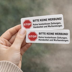 Könnte beinhalten: Zwei weiße rechteckige Schilder mit dem Text "BITTE KEINE WERBUNG!" und "STOP" in einem roten Achteck. Die Schilder enthalten auch deutschen Text über unerwünschte Werbung. Die Schilder werden in einer Hand gehalten.