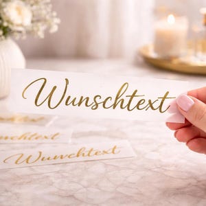 Könnte beinhalten: Eine Hand hält eine weiße rechteckige Karte mit dem goldenen Schriftzug "Wunschtext". Mehrere ähnliche Karten liegen auf einer Marmoroberfläche. Der Hintergrund umfasst eine weiße Vase mit Blumen, eine Kerze und eine Parfümflasche.