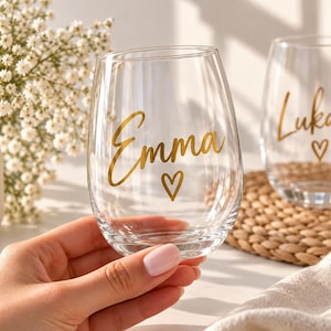 Könnte beinhalten: Zwei klare, stiellose Weingläser aus Glas. Das vordere Glas zeigt den Namen Emma in goldener Schreibschrift mit einem kleinen goldenen Herz darunter. Das zweite Glas im Hintergrund zeigt den Namen Lukas in goldener Schreibschrift mit einem Herz.