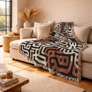 May include: A woven throw blanket with a geometric pattern in black, cream, and rust colors. The blanket is draped over a beige sofa and features the name "Charlotte" in white text. The fringe adds a decorative touch.