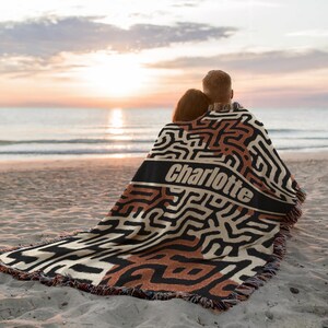 May include: A woven blanket with a geometric pattern in black, cream, and brown, personalized with the name "Charlotte" in white. The blanket has a fringed edge and is draped over two people on a sandy beach at sunset.