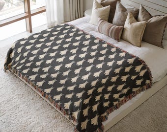 May include: A dark gray and cream-colored throw blanket with a repeating geometric pattern, fringed edges, and a textured appearance. The blanket is displayed on a bed with pillows.
