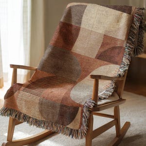 May include: A woven throw blanket with a geometric pattern in shades of brown, tan, and beige is draped over a wooden rocking chair. The blanket has a fringed edge. The chair is made of light-colored wood.