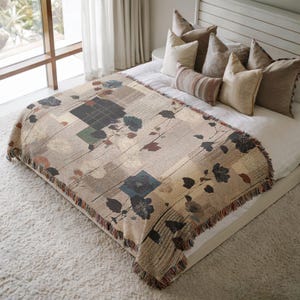 May include: A beige throw blanket with a floral pattern and fringed edges is draped over a bed. The blanket features squares and rectangles with floral and leaf designs in shades of blue, brown, and beige. The bed is made with white sheets and several pillows.