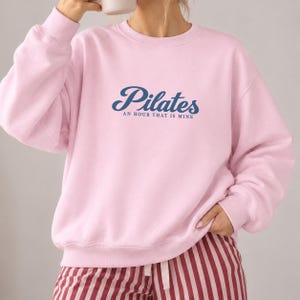 May include: A light pink crewneck sweatshirt featuring the dark blue text "Pilates" in a cursive font, with the smaller text "AN HOUR THAT IS MINE" printed directly underneath in a simple, clean, sans-serif typeface.