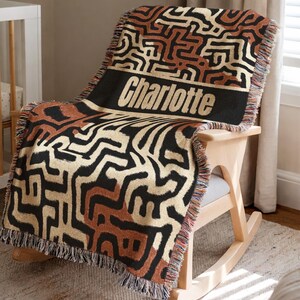 May include: A woven throw blanket with a geometric pattern in black, tan, and brown is draped over a wooden rocking chair. The blanket has a black band with the name "Charlotte" in white letters. The blanket has a fringed edge.