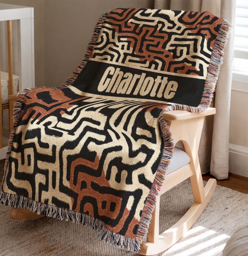 May include: A woven throw blanket with a geometric pattern in black, tan, and brown is draped over a wooden rocking chair. The blanket has a black band with the name "Charlotte" in white letters. The blanket has a fringed edge.