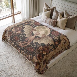 May include: A woven throw blanket featuring a dragon design with a full moon in the background. The blanket has a dark background with floral accents and a fringed edge. The dragon is depicted in shades of brown and tan.