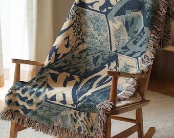 May include: A woven throw blanket draped over a wooden rocking chair. The blanket features a geometric pattern in shades of blue, beige, and green, with fringed edges. The chair is made of light-colored wood and is positioned in front of a window.