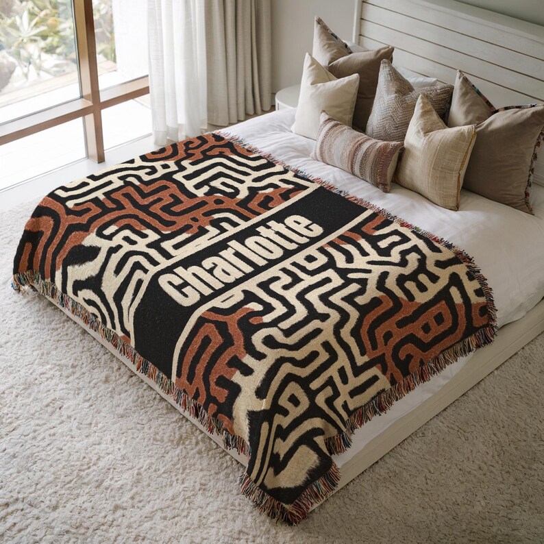May include: A woven throw blanket with a geometric pattern in black, brown, and cream. The blanket features the name "Charlotte" in white block letters on a black stripe. The blanket is fringed and placed on a bed with pillows.