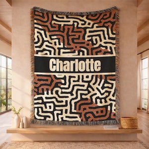May include: A woven throw blanket with a geometric pattern in black, brown, and cream colors. The blanket has a black band with the name "Charlotte" in white letters. The blanket is fringed on the edges.