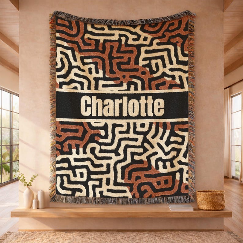 May include: A woven throw blanket with a geometric pattern in black, brown, and cream colors. The blanket has a black band with the name "Charlotte" in white letters. The blanket is fringed on the edges.