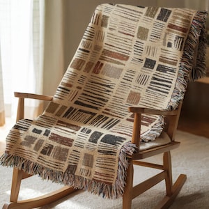 May include: A beige throw blanket with a geometric pattern of brown, black, and gray rectangles and lines, draped over a wooden rocking chair. The blanket has a fringed edge with multiple colors.