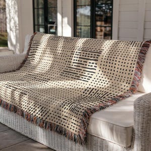 May include: Cream-colored woven throw blanket with a black geometric pattern of dots and lines. The blanket has a fringed edge with orange, brown, and blue accents. The throw is draped over a white wicker sofa.