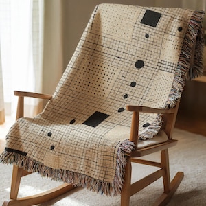 May include: A beige throw blanket with a geometric pattern of black squares, dots, and lines is draped over a wooden rocking chair. The blanket has a fringed edge with multiple colors. The chair is made of light-colored wood.
