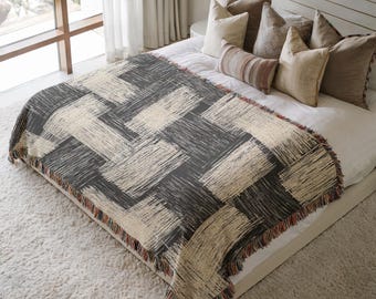 May include: A woven throw blanket with a geometric pattern in shades of gray and beige. The blanket has a fringed edge with red, brown, and beige threads. The blanket is displayed on a bed with pillows.