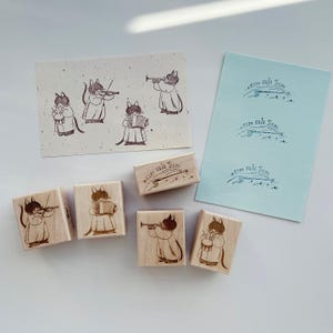 Peut inclure: Ensemble de tampons en bois représentant des chats musiciens jouant de divers instruments, dont un violon, un accordéon, une trompette et une batterie. Les tampons sont accompagnés d'images estampées sur du papier bleu clair et beige. Le texte "sim sala bim" est également estampé.