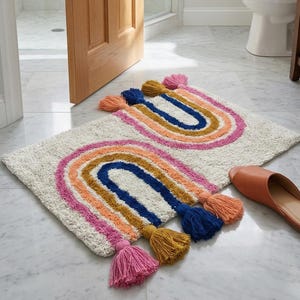 Puede incluir: Dos alfombras de baño rectangulares de color blanquecino con arcos de colores arcoíris y borlas. Los arcos son en tonos rosa, naranja, dorado y azul. Un par de zapatillas de cuero marrón están cerca. Las alfombras están sobre un suelo de mármol.