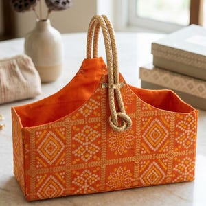 Può includere: Un cestino in tessuto arancione con un motivo geometrico giallo e bianco. Il cestino ha un manico intrecciato e un interno arancione abbinato. Il cestino è su un tavolo di marmo.